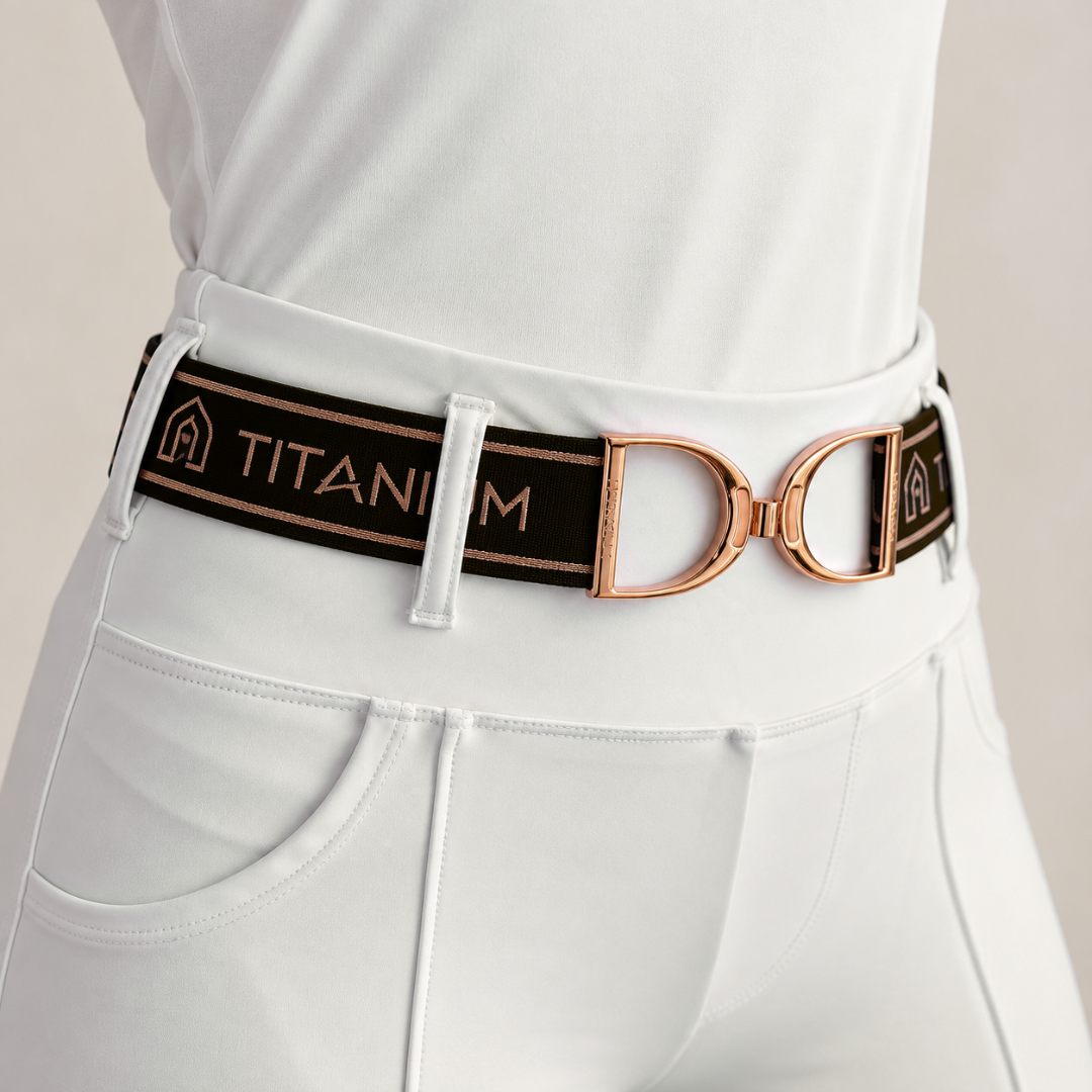 A close-up of a black elastic belt with rose gold hardware, worn by a model in white riding pants. The belt features the text 'TITANIUM' in rose gold with a small logo on the left, and a large rose gold stirrup buckle.