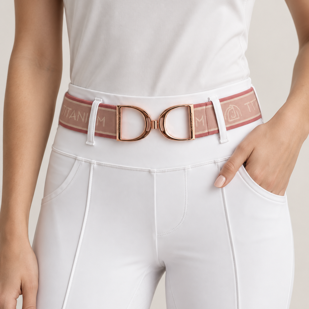 A close-up of a model wearing white riding tights and a pink and rose gold elastic belt with a large rose gold stirrup buckle. The belt features the brand name 'TITANIUM' embroidered on the elastic.
